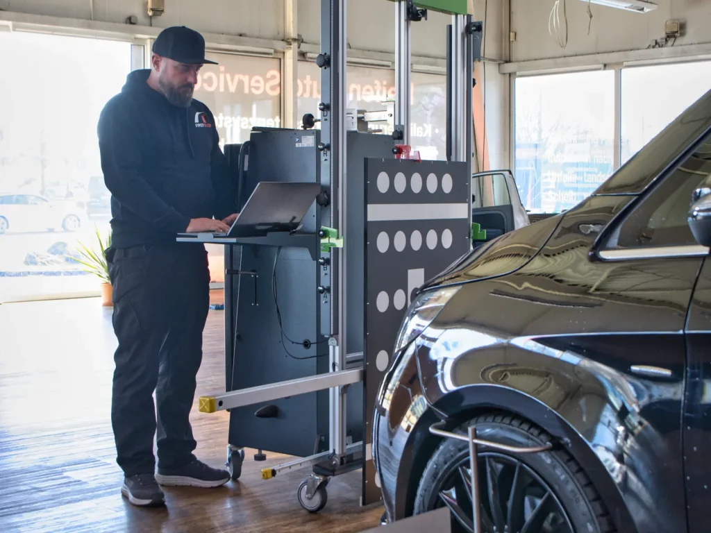 Mitarbeiter + arbeitet an einem Computer neben einem modernen Gerät + FIRSTSTOP Schwerin Werkstattumgebung, sichtbar ist der vordere Teil eines schwarzen Fahrzeugs.
