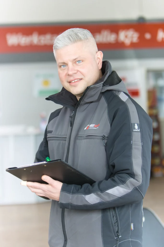 Mitarbeiter in Werkstattkleidung hält ein Klemmbrett, Hintergrund mit Werkschild. Zusammenhang mit FIRSTSTOP Schwerin.